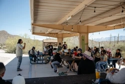 Students listen intently to an educator who is facing them and providing a lesson underneath an outside ramada.