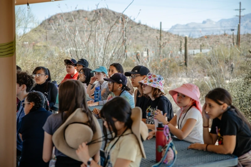 a group of students are seated at outside tables under a ramada during an orientation meeting at Camp Cooper