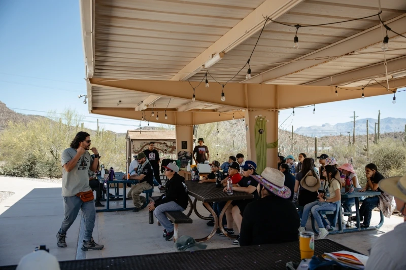a group of students are seated at outside tables under a ramada during an orientation meeting at Camp Cooper