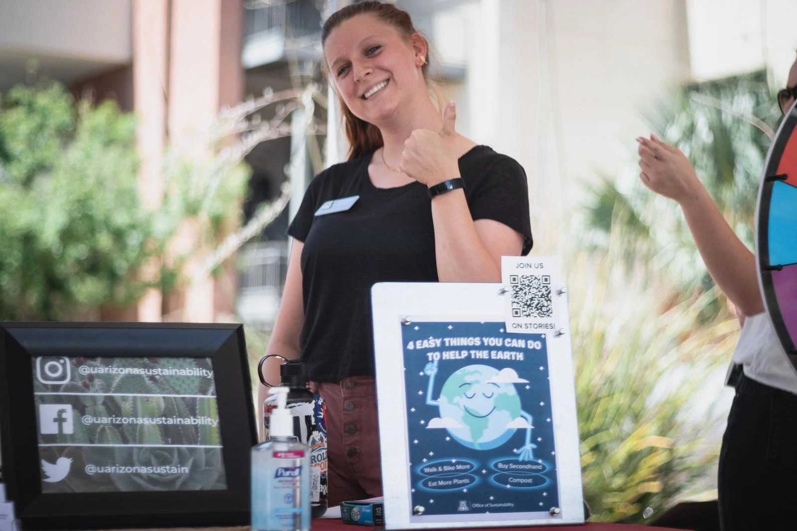 Campus Sustainability Fund committee member, Madison Gerdes, at the Office of Sustainability table in front of a poster listing 4 easy things to do to help the Earth - Walk & Bike More, Buy Secondhand, Eat More Plants, and Compost. The image also includes all the social links of Office of Sustainability.