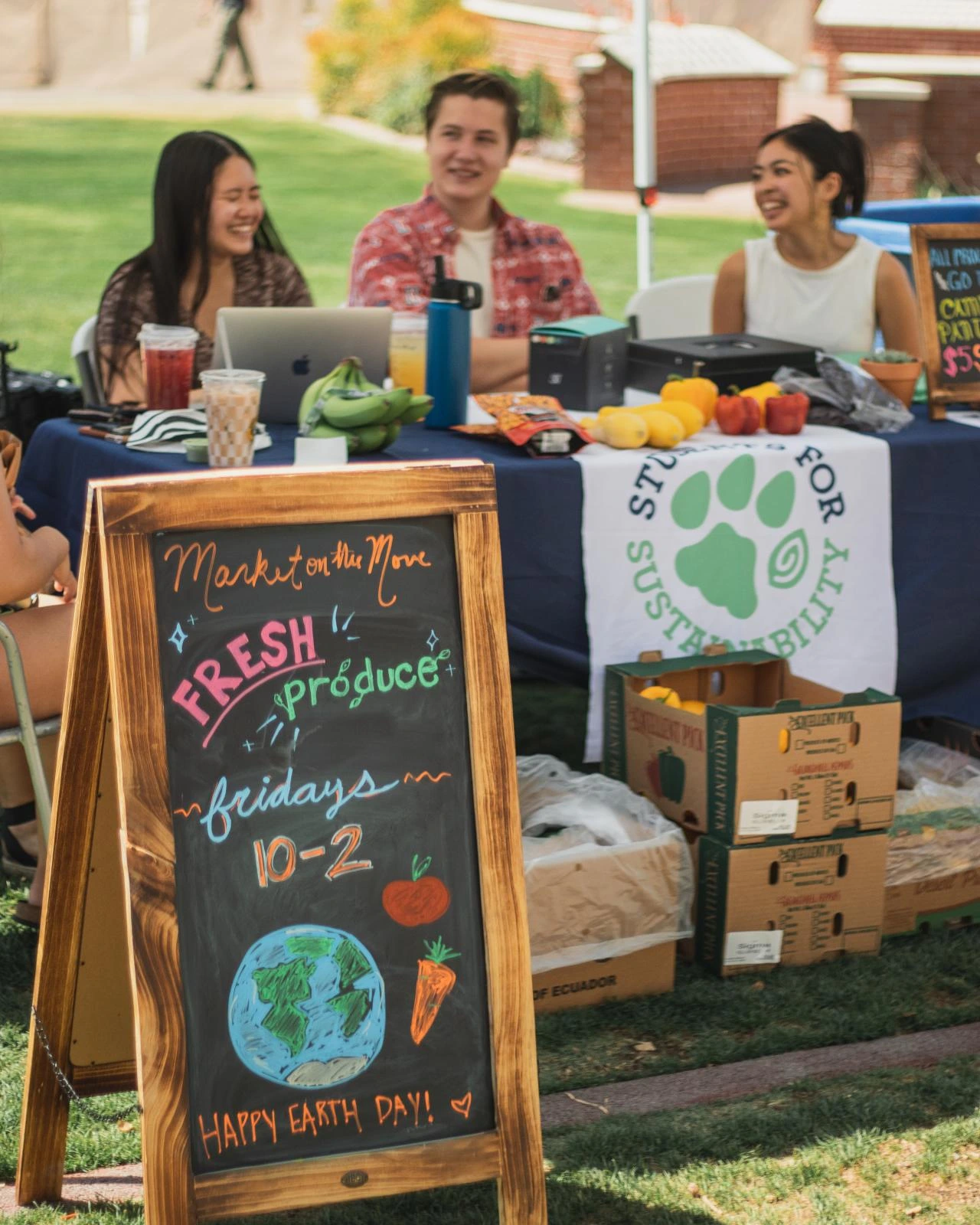 A sign reads "Market on the Move: Fresh Produce, Fridays 10-2" in front of a few boxes of produce. 