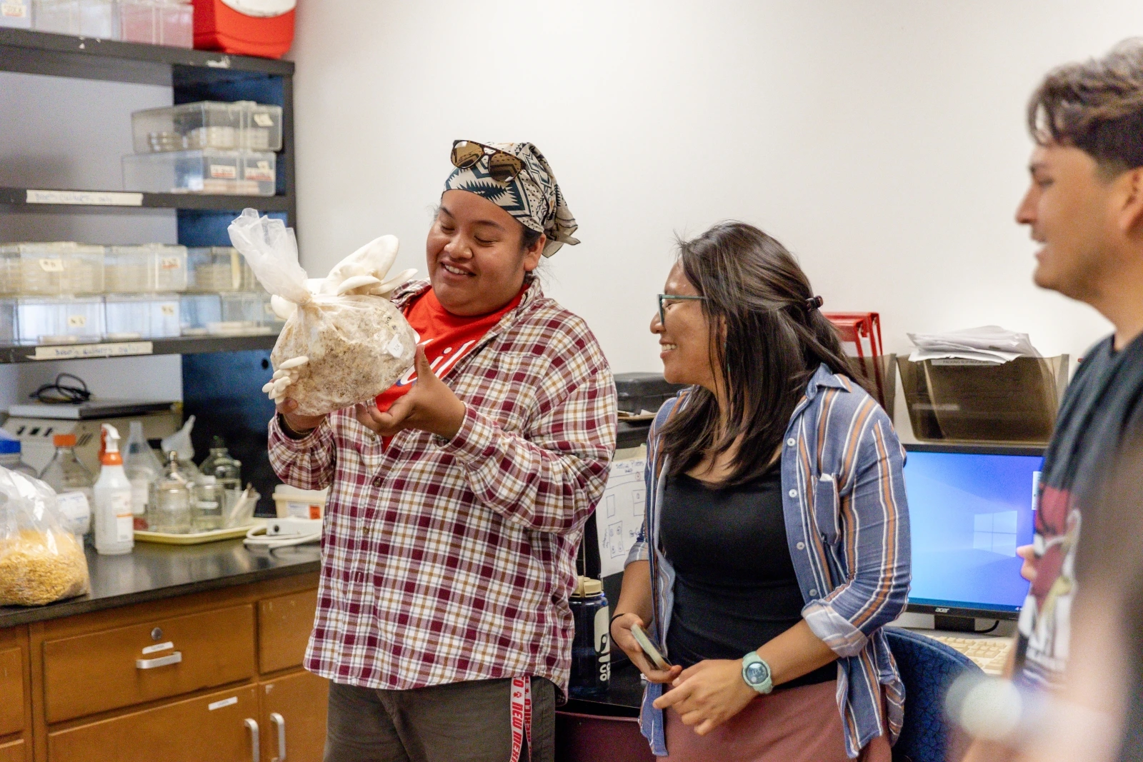 Three people are standing next to each other while all looking at the person to the left. The person on the left is holding a bag with a large white mushroom inside of it.
