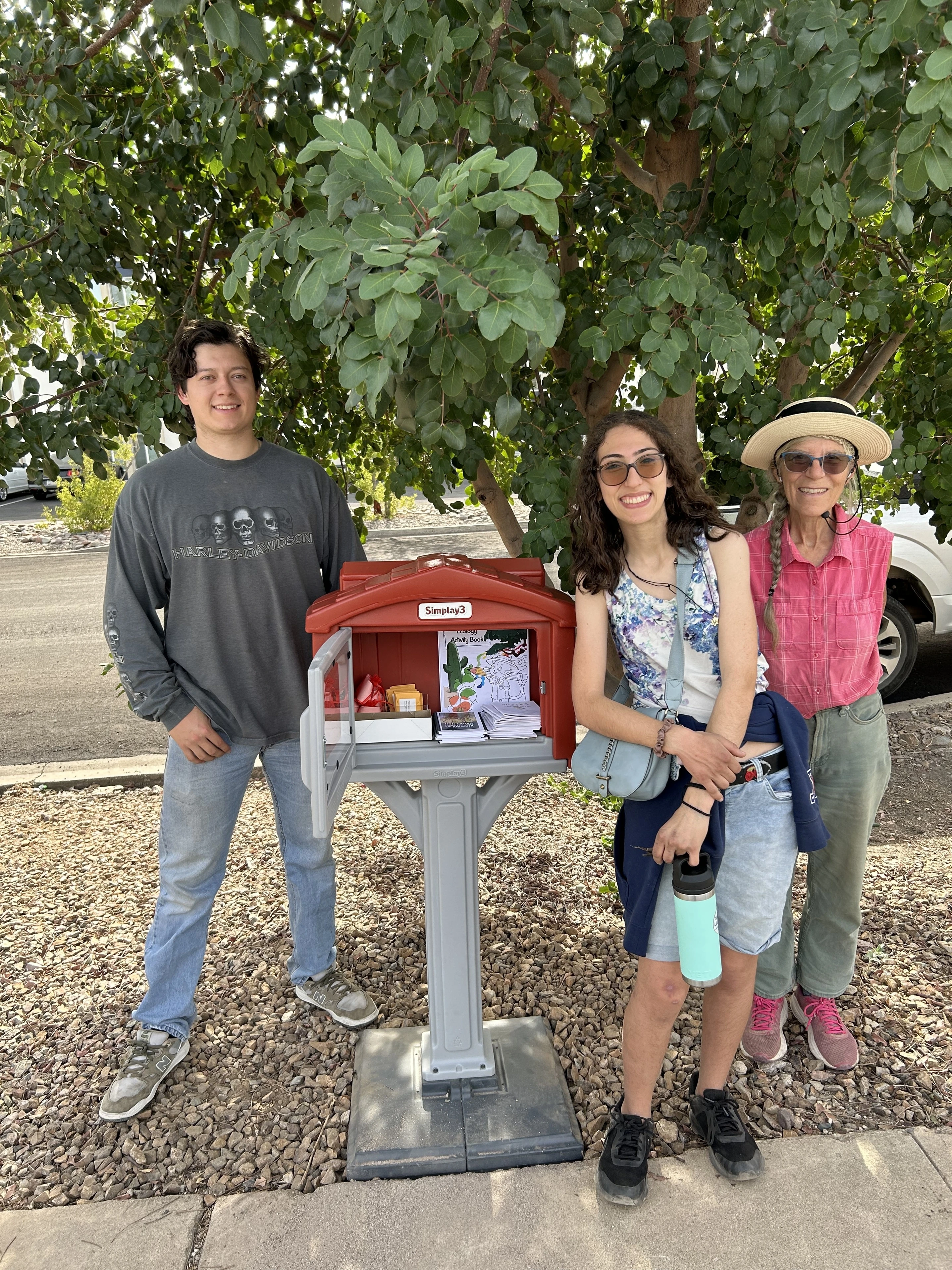 Three people stand under a tree next to a small seed library