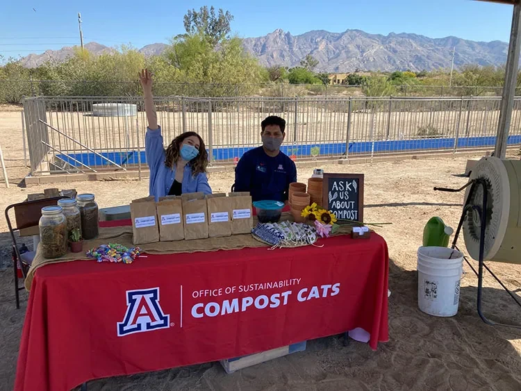 Two Compost Cats student employees sit at a table displaying the Compost Cats logo and outreach materials.