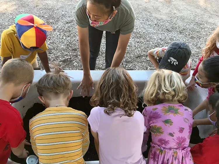 A group of children gather around a rectangular container filled with soil, participating in an outdoor gardening activity. An adult stands in the middle, leaning over the container to guide or instruct them. The ground is covered with wood chips.