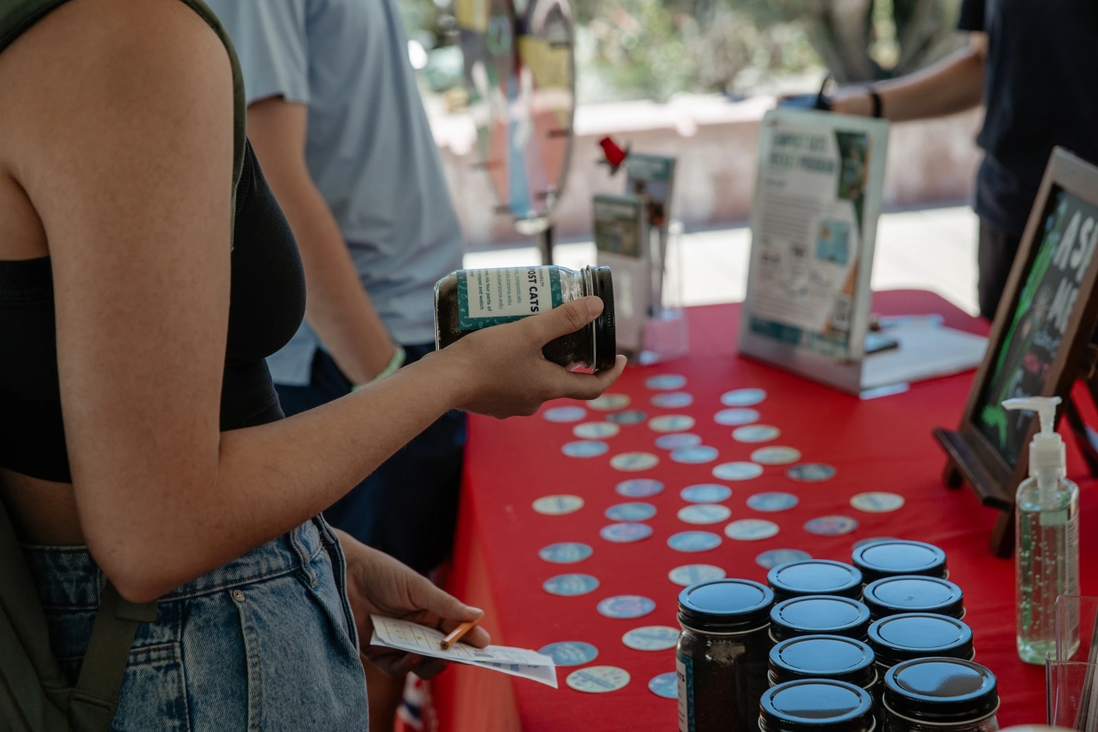 A table covered in red cloth that is filled with stickers, jars filled with compost, and informational cards. A person is standing near the table holding a compost jar.