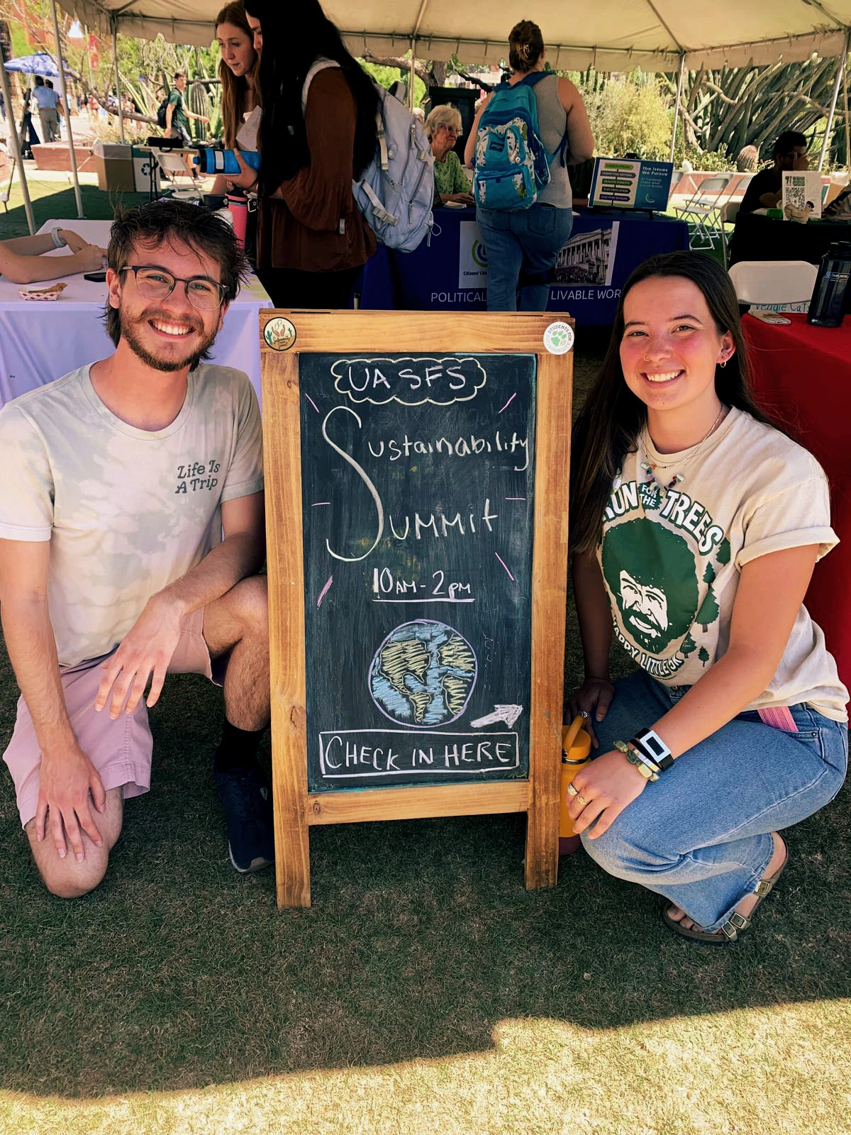 Two people are squatting down next to a chalkboard sign that reads "UA SFS, Sustainability Summit, 10am - 2pm, Check in here".