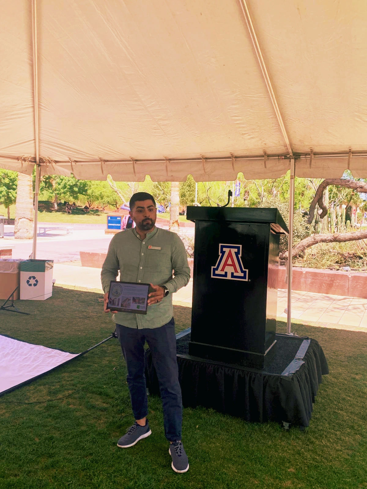A person stands in front of a podium talking to a group of people under a white tent.
