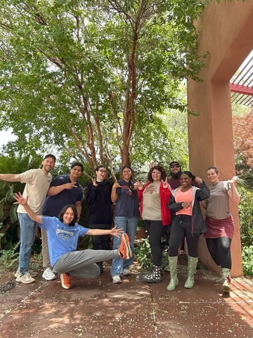 A group of nine people smile and pose together in front of a tree