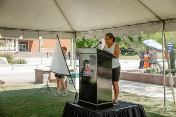 A person standing on a podium speaking to a group of people. The podium is under a white tent.