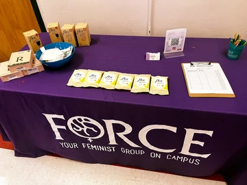 A table covered in a purple cloth with the FORCE logo. On top of the table are various boxes of menstrual cups and wipes. 