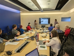 Two students stand near a screen and are presenting to a large team of people seated at a conference table.