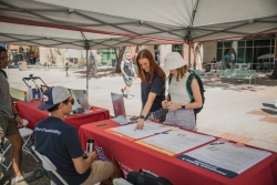 Two people standing near a red table looking at 3 big posters laid out on the table. While another person is sitting on the opposite side of the table talking to them.