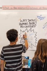 Two people standing in front of a white board where a piece of paper is taped to the whiteboard. They are writing on the paper. 