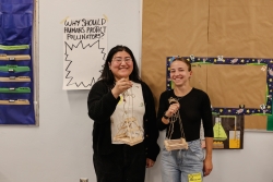 Two people standing next to each other, smiling, and holding up structures made out of wooden popsicle sticks.