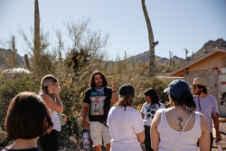 A group of people standing outside in a desert area, looking at a person talk to them.