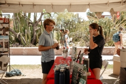 Two people talking to each other with a red table in between them. One person is holding a jar filled with compost.