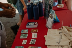 A table covered in red cloth that is filled with stickers, water bottles, and tote bags. A person's hand is shown holding stickers.