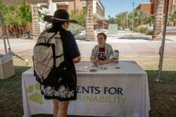 A person talking to another person that is sitting behind a table covered in white cloth. "Students for Sustainability" is written on the white cloth.