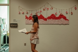 A person holding a paper talking while standing in front of an art installation on a wall behind them.