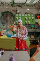 A person standing in front of a table and talking to an group of people with one hand raised.