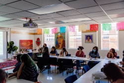 A room with desks laid out in a circle. Multiple people are sitting and listening to a presentor.