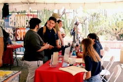 People standing next to a table covered in outreach material. They are holding pamphlets and talking to the people sitting behind the table.