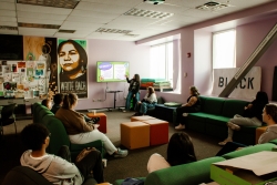 A group of people sitting around a common area looking at a tv. A person next to the tv has their hand raised to point at the screen.