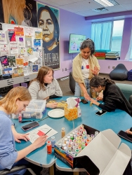 A group of people sitting at a table painting. One person is standing while getting their nails painted by another person sitting next to them. 