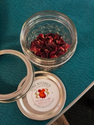 A small glass jar has rose petals inside it. The lid of the jar is laid out next to the glass jar with a circular sticker on top of the lid.