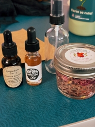 Two droppers filled with different oils and herbs are next to each other. There is a small glass jar next to the two droppers that is filled with water and rose petals. A glass spray bottle is standing behind the jar and droppers.