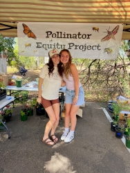 Two people hugging and smiling in front of a hanging sign that reads "Pollinator Equity Project".