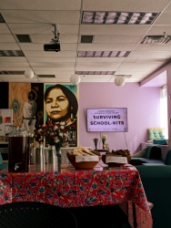 A room with purple walls and various large art pieces are hung on the wall. There is a table covered in red cloth with a jug of tea, cups, and cookies on top of the table. At the far wall behind the table, there is a television.