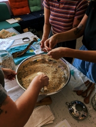 Three people reaching into a large metal bowl filled with various herbs. The bowl is set on a table with white cloth and various appliances like wooden spoons and cloth surround the bowl.. 