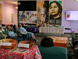 A room with purple walls and various large art pieces are hung on the wall. There are multiple colorful couches laid out in an open circle with people sitting on them. The people have a small bag of herbs covering their eyes.