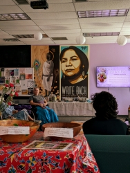 A room with purple walls and various large art pieces are hung on the wall. There is a table covered in red cloth with a jug of tea, cups, and cookies on top of the table. At the far wall behind the table, there is a television and a person is presenting next to the tv to a group of people.