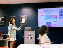 A person standing in front of a group of people pointing at a television and talking to the group.