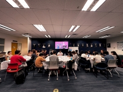 A large group of people all sitting in three tables making clay cherry blossoms.