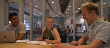 Three students laughing while talking to each other at a table with laptops and notebooks in front of them with windows and the Canyon Cafe in ENR2 in the background. 