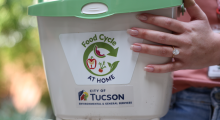 A very large diamond engagement ring sparkles on a hand holding a bucket for collecting food scraps with Food Cycle at Home and City of Tucson labels on it