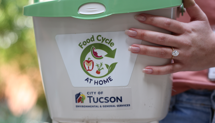 A very large diamond engagement ring sparkles on a hand holding a bucket for collecting food scraps with Food Cycle at Home and City of Tucson labels on it