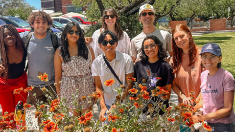 A group of people standing next to each other and smiling behind a flower bush.