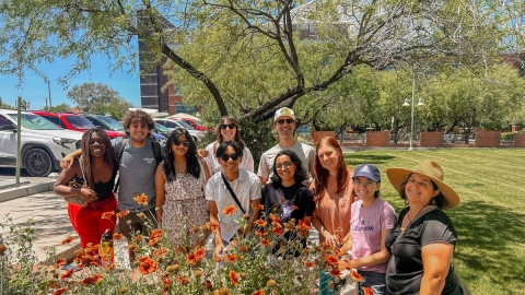 A group of people stand behind a large flowering planter that is outside between a parking lot and grass lawn