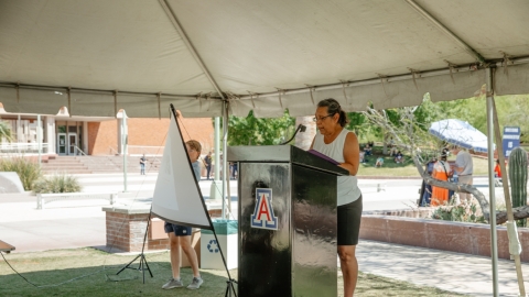 A person standing on a podium speaking to a group of people. The podium is under a white tent.