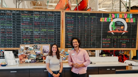 Two people standing next to each other behind a front desk and in front of a large chalk board, smiling for a photo.