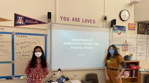 Two classroom educators pose in front of their lesson board. 