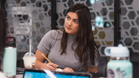A young woman with long dark hair wearing a gray short-sleeve turtleneck sweater sits at a table, attentively listening and taking notes with a pencil. The background features a modern office or classroom setting with a hexagonal-patterned glass partition.