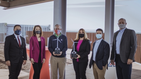 A group of six professionally dressed individuals stand side by side outdoors under a covered structure, all wearing face masks. Two individuals in the center are holding small wind turbine-shaped awards.